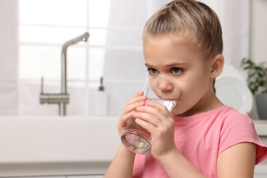 Photo of Cute little girl drinking fresh water from glass in kitchen