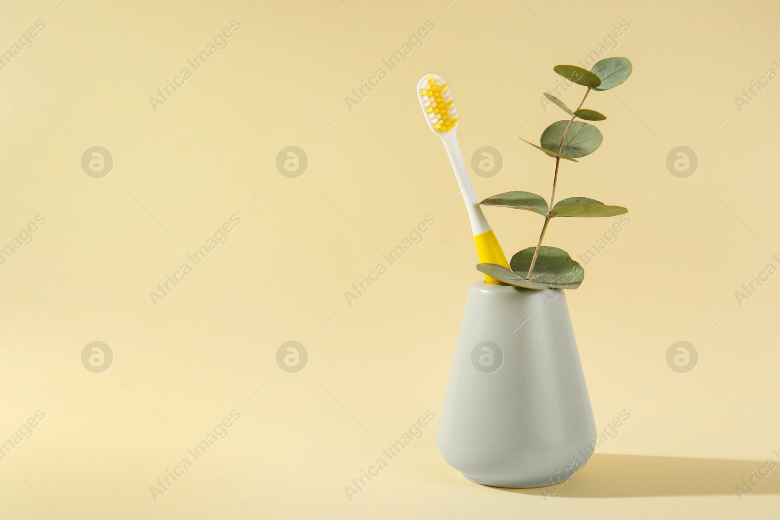 Photo of Plastic toothbrush and eucalyptus branch in holder on pale yellow background, space for text