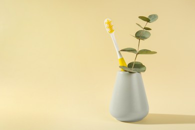 Plastic toothbrush and eucalyptus branch in holder on pale yellow background, space for text