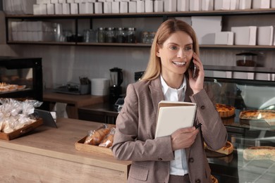 Photo of Happy business owner with notebook and pen talking on phone in bakery shop