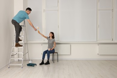 Photo of Young woman giving roller to man on stepladder indoors. Room renovation