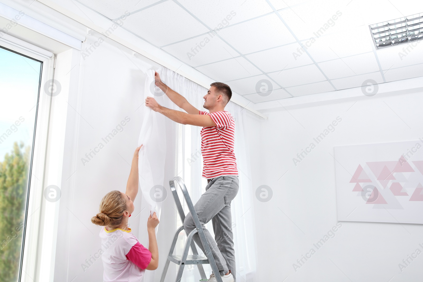 Photo of Young couple hanging window curtain indoors. Interior decor element