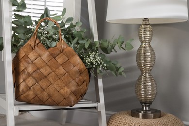 Photo of Stylish straw bag with beautiful bouquet and lamp indoors