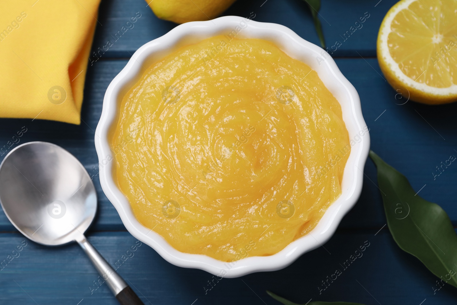 Photo of Delicious lemon curd in bowl, fresh citrus fruits and spoon on blue wooden table, flat lay