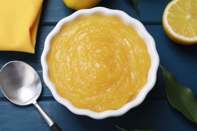 Delicious lemon curd in bowl, fresh citrus fruits and spoon on blue wooden table, flat lay