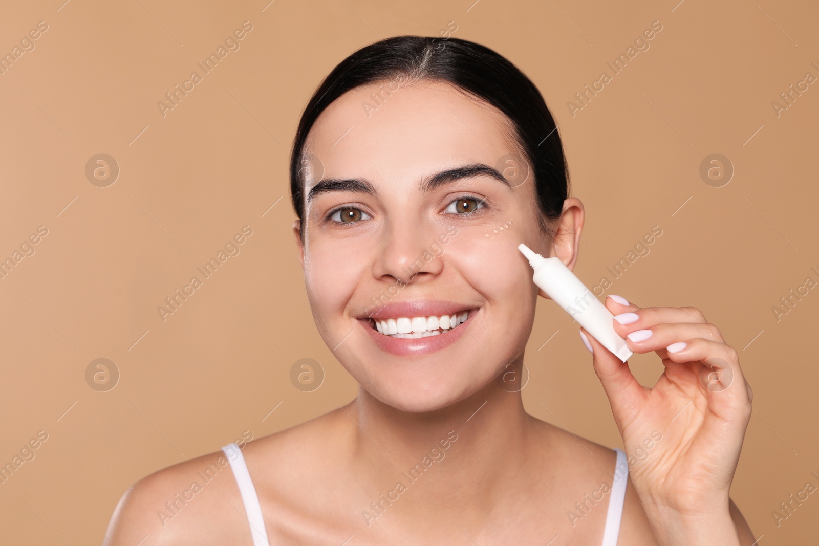 Photo of Beautiful young woman applying gel on skin under eye against beige background