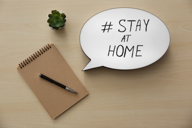 Notepad, houseplant and speech bubble with hashtag STAY AT HOME on wooden background, flat lay. Message to promote self-isolation during COVID‑19 pandemic