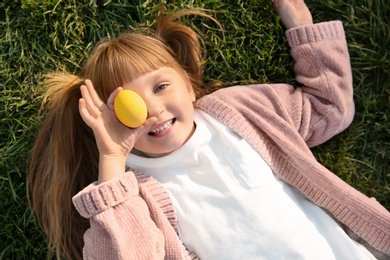 Photo of Cute little girl holding Easter egg while lying on green grass, top view