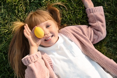 Cute little girl holding Easter egg while lying on green grass, top view