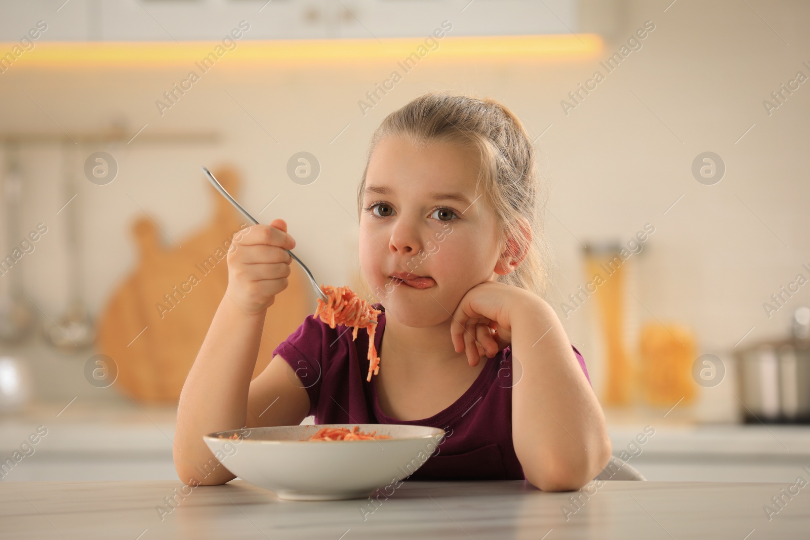 Photo of Cute little girl eating tasty pasta at table in kitchen