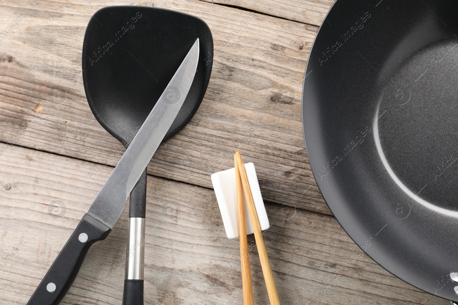 Photo of Black metal wok, chopsticks, knife and spatula on wooden table, flat lay