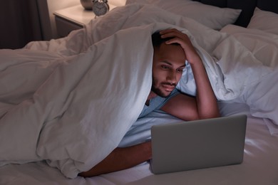 Photo of Young man using laptop under blanket in bed at night. Internet addiction