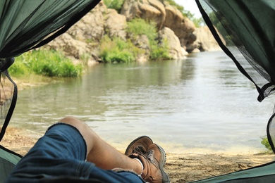Photo of Young man resting in camping tent on riverbank, view from inside