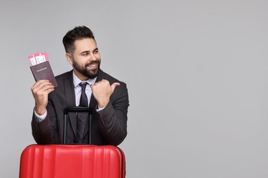 Photo of Happy businessman with passport, tickets and suitcase pointing at something on grey background. Space for text