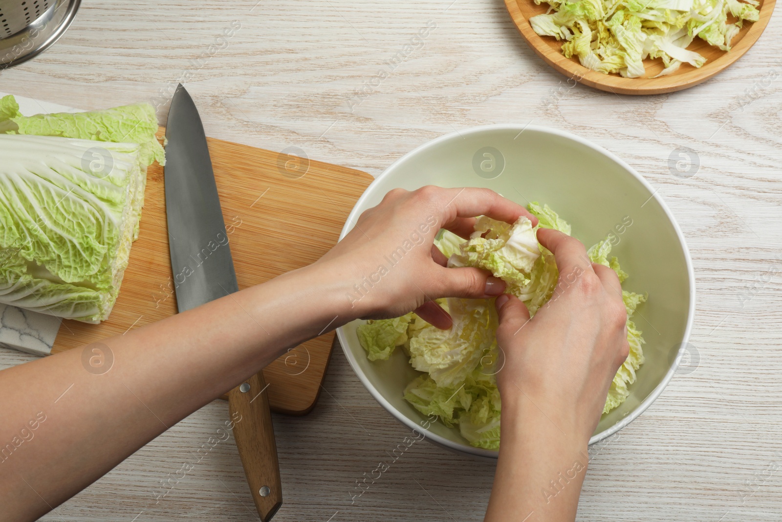 Photo of Woman putting cut Chinese cabbage into bowl at white wooden kitchen table, top view