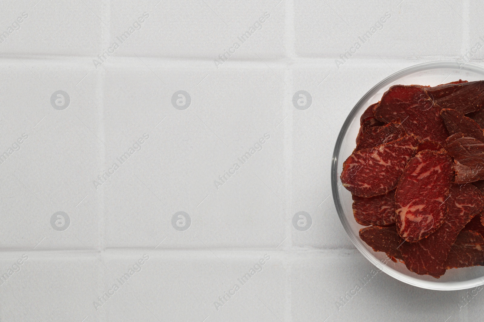 Photo of Bowl with pieces of delicious beef jerky on white tiled table, top view. Space for text