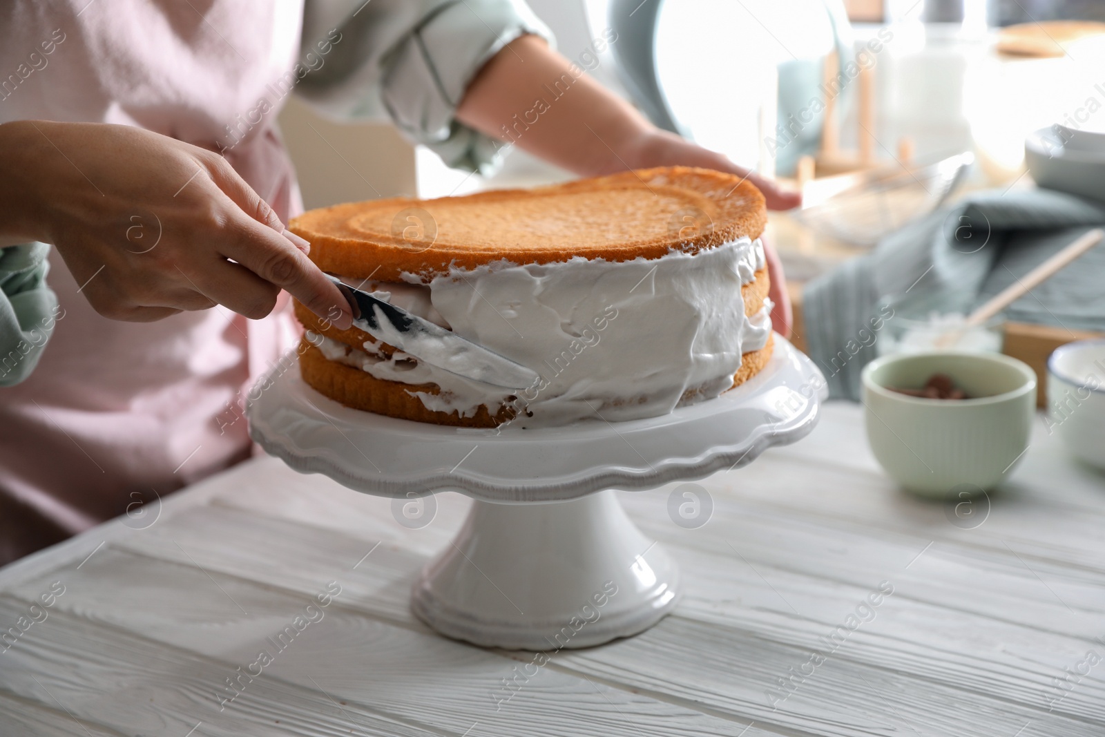 Photo of Woman smearing sides of sponge cake with cream at white wooden table, closeup