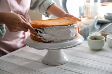 Photo of Woman smearing sides of sponge cake with cream at white wooden table, closeup