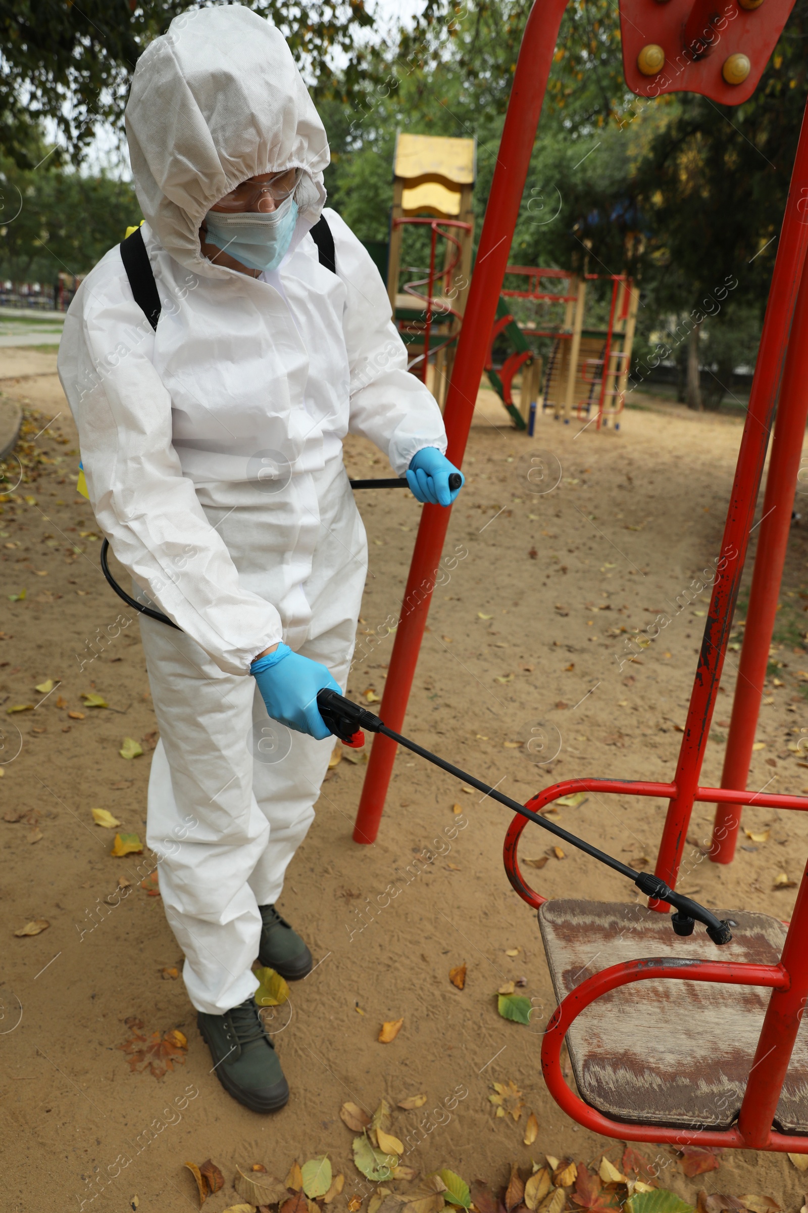 Photo of Woman wearing chemical protective suit with disinfectant sprayer on playground. Preventive measure during coronavirus pandemic