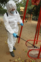 Photo of Woman wearing chemical protective suit with disinfectant sprayer on playground. Preventive measure during coronavirus pandemic