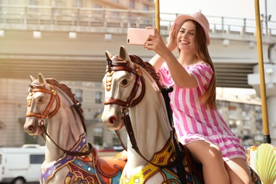Photo of Young pretty woman taking selfie on carousel in amusement park