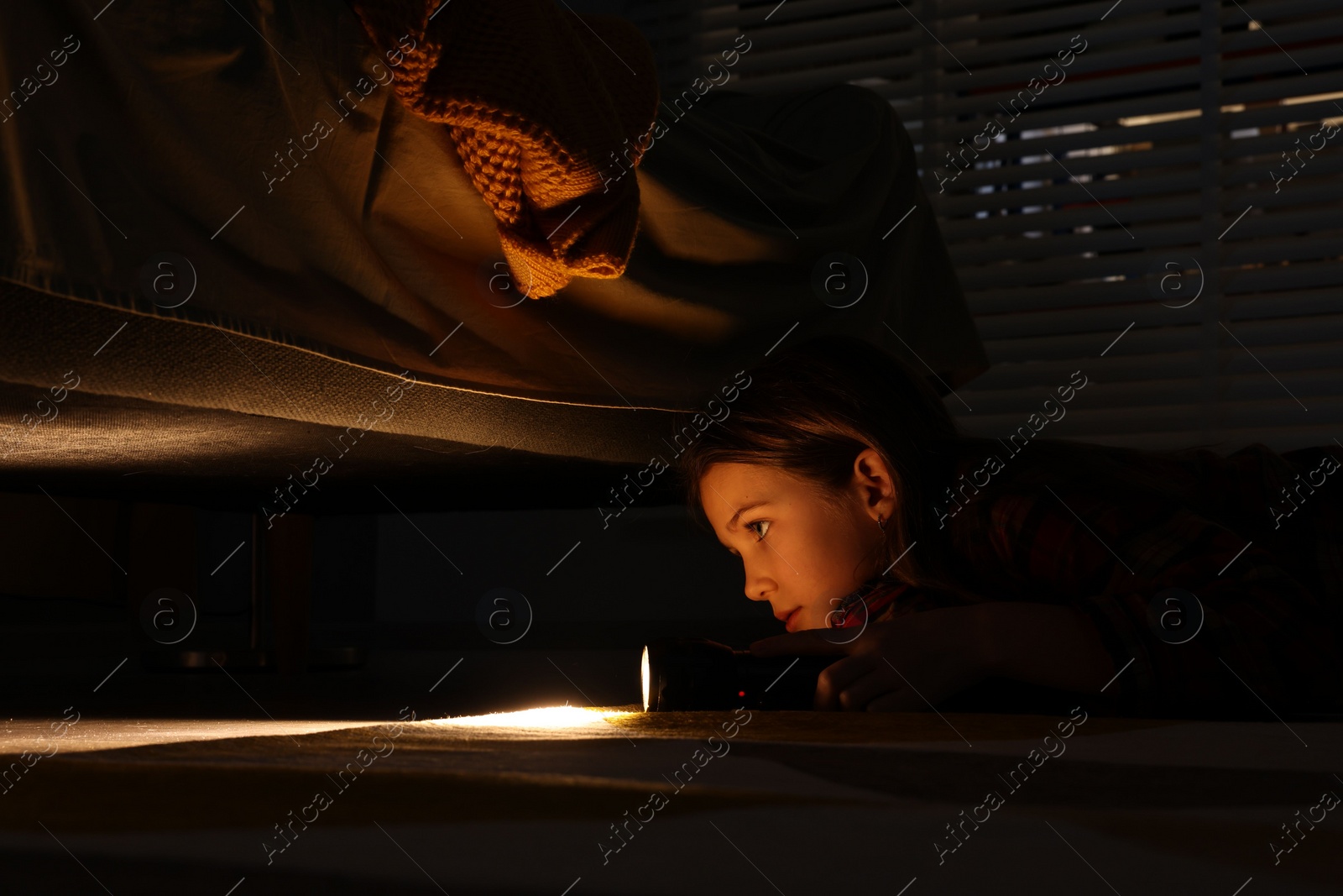 Photo of Little girl with flashlight looking for monster under bed at night