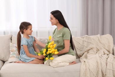 Happy woman and her cute daughter with bouquet of yellow tulips on sofa at home. Mother's day celebration