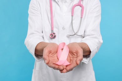 Photo of Doctor with stethoscope holding pink ribbon on light blue background, closeup. Breast cancer awareness