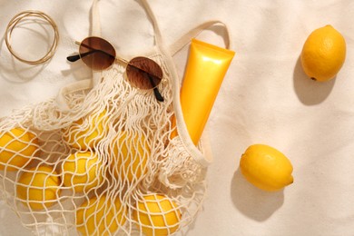 Photo of String bag with sunglasses, lemons and sunscreen on sand, flat lay