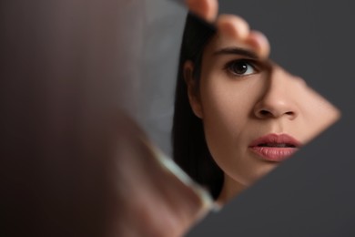 Young woman looking at herself in shard of broken mirror on grey background, closeup