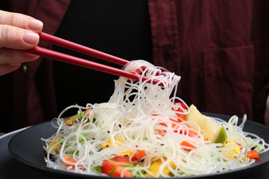 Photo of Stir-fry. Woman with chopsticks eating tasty rice noodles with meat and vegetables at table, closeup