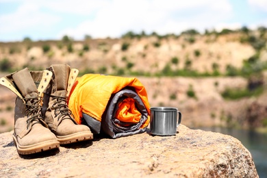Photo of Sleeping bag, cup and boots on cliff near lake. Space for text