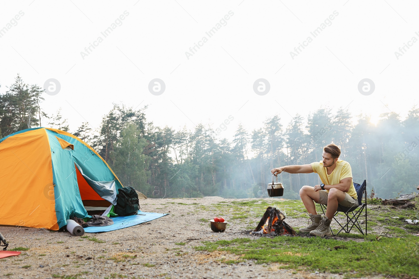 Photo of Young man holding kettle over bonfire near camping tent outdoors