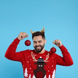 Photo of Happy young man in Christmas sweater and reindeer headband holding festive baubles on light blue background
