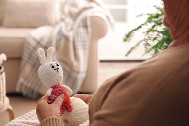 Photo of Elderly woman with knitted bunny at home, closeup. Creative hobby
