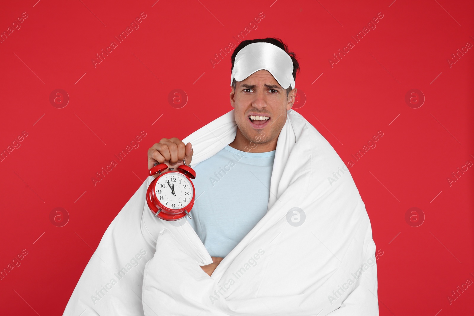 Photo of Emotional man in sleeping mask wrapped with blanket holding alarm clock on red background