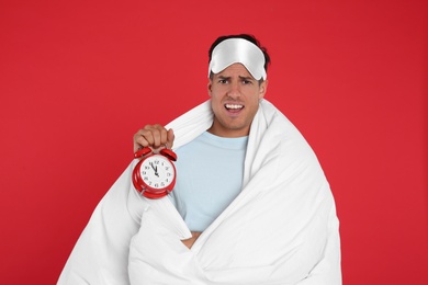 Photo of Emotional man in sleeping mask wrapped with blanket holding alarm clock on red background