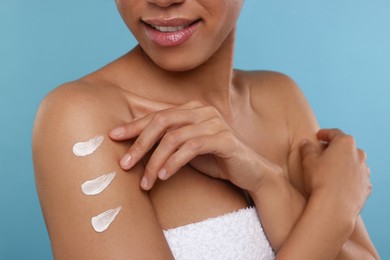 Young woman applying body cream onto arm on light blue background, closeup