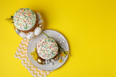 Traditional Easter cakes with sprinkles, painted eggs and beautiful spring flowers on yellow background, flat lay. Space for text