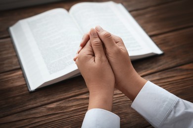 Woman holding hands clasped while praying at wooden table with Bible, closeup