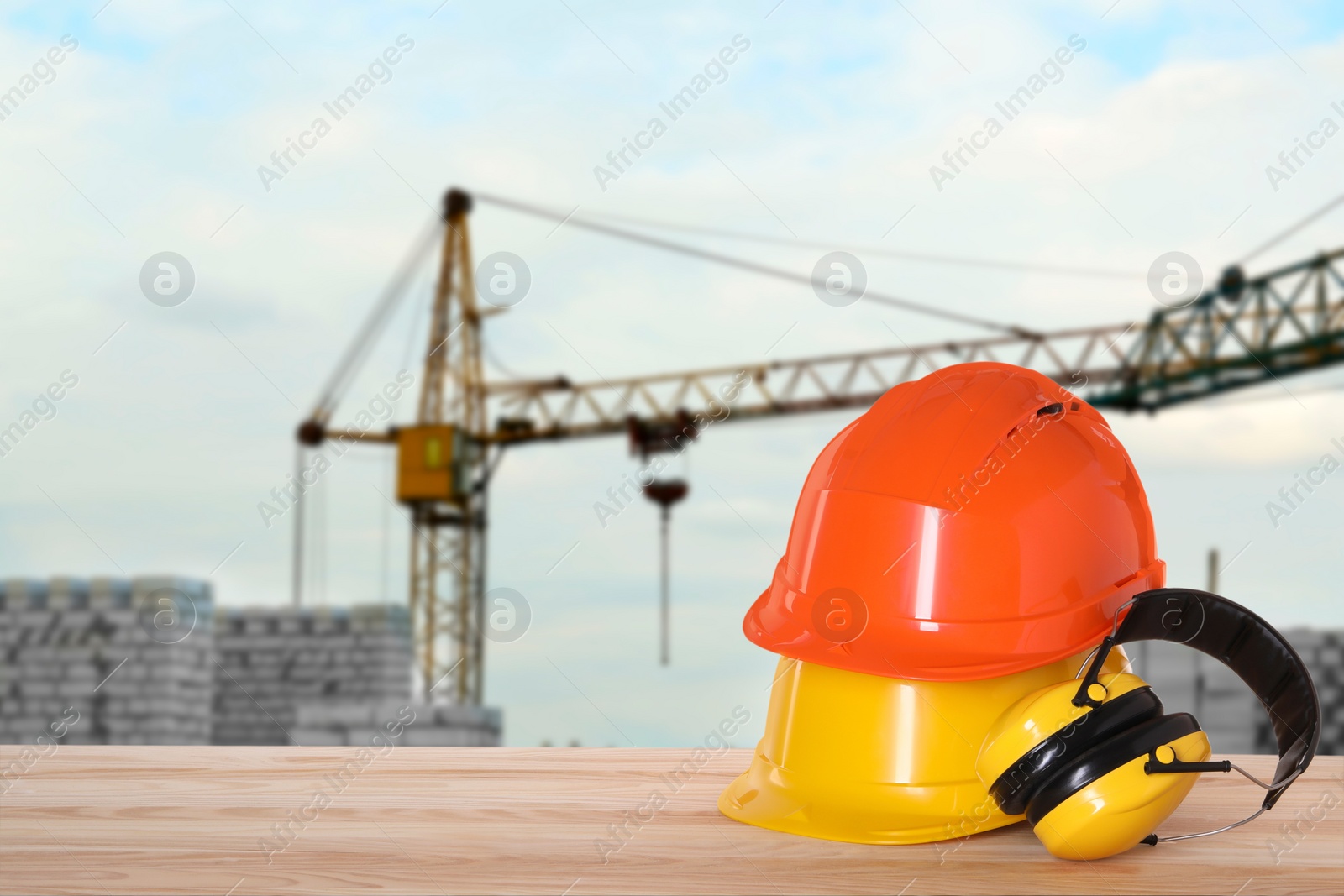 Image of Safety equipment. Hard hats and protective headphones on wooden surface near construction site with tower crane outdoors, space for text