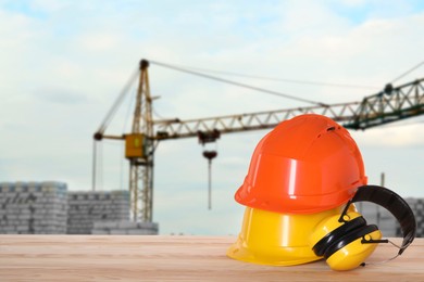 Image of Safety equipment. Hard hats and protective headphones on wooden surface near construction site with tower crane outdoors, space for text