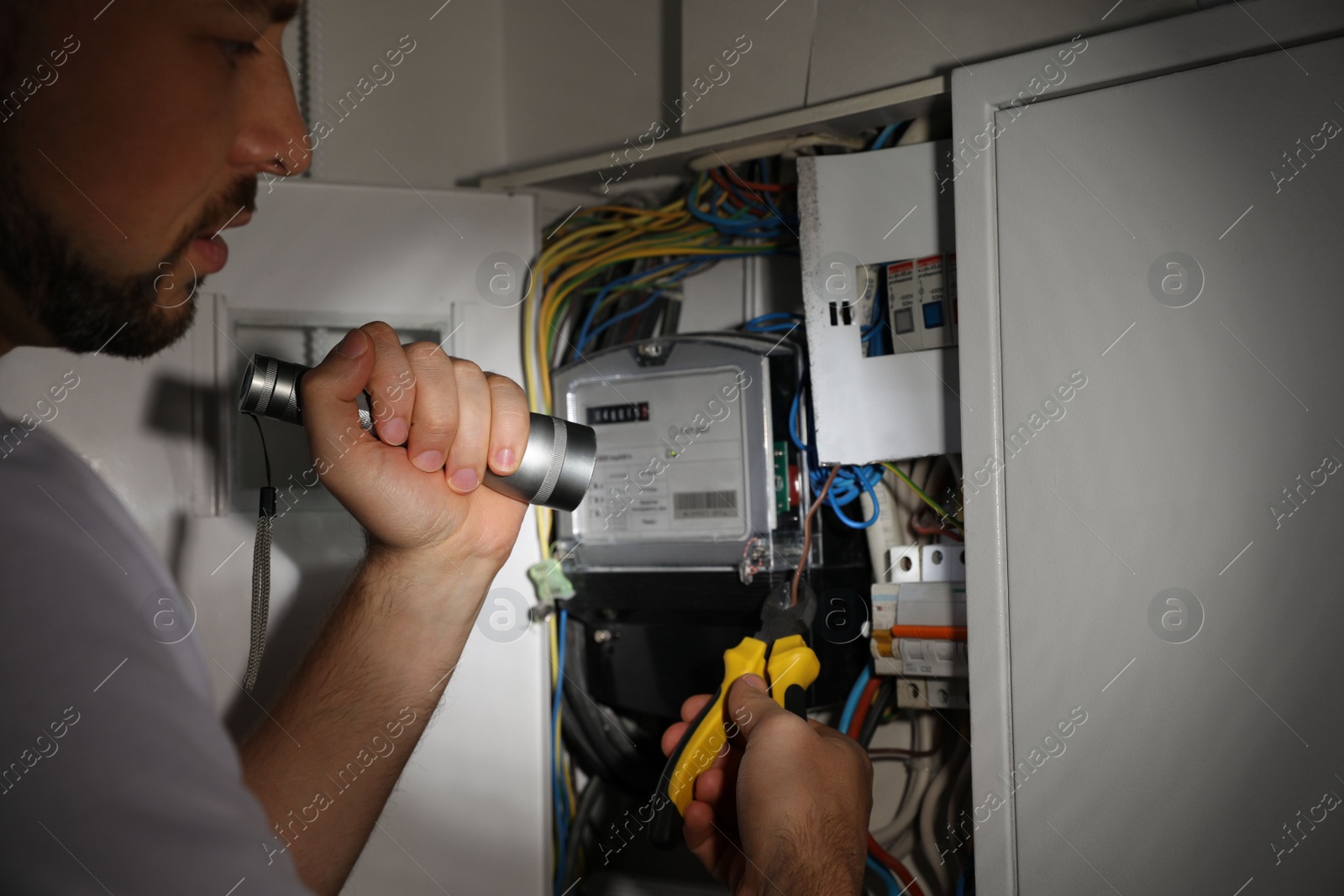 Photo of Electrician with flashlight fixing electric panel indoors, closeup