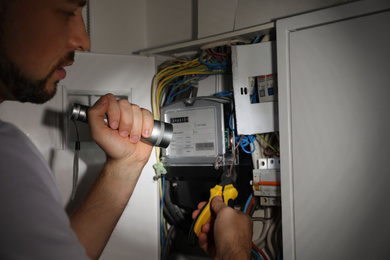 Electrician with flashlight fixing electric panel indoors, closeup