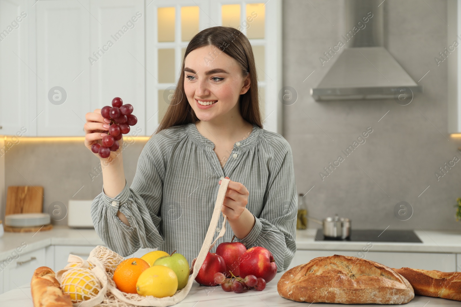 Photo of Woman taking grapes out from string bag at light marble table