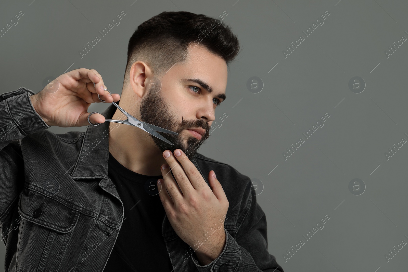 Photo of Handsome young man trimming beard with scissors on grey background. Space for text