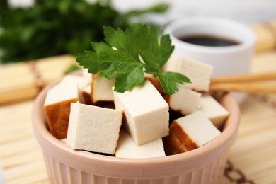 Photo of Bowl of smoked tofu cubes, soy sauce and parsley on bamboo mat, closeup