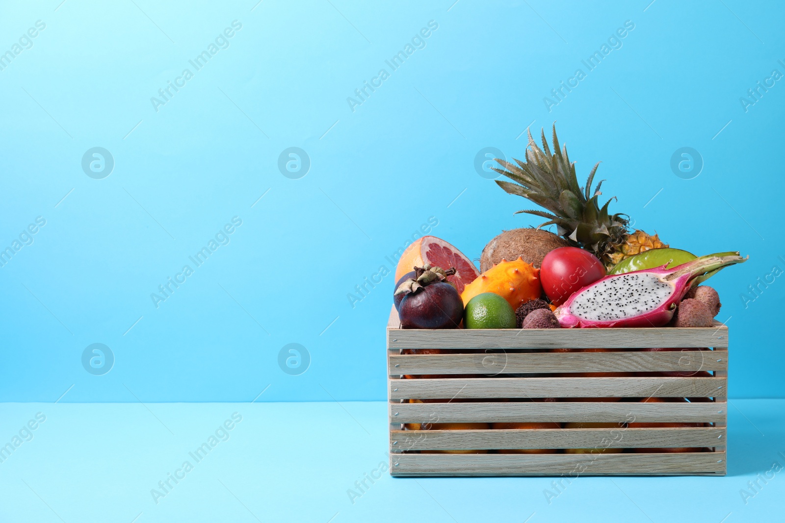 Photo of Different tropical fruits in wooden box on light blue background. Space for text