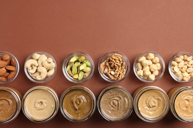 Photo of Many tasty nut butters in jars and nuts on brown table, flat lay. Space for text