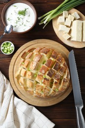 Photo of Freshly baked bread with tofu cheese, green onions, sauce and knife on wooden table, flat lay